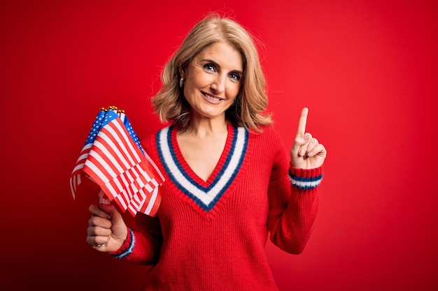 Free photo middle age blonde patriotic woman holding united states flags celebrating independence day surprised with an idea or question pointing finger with happy face number one