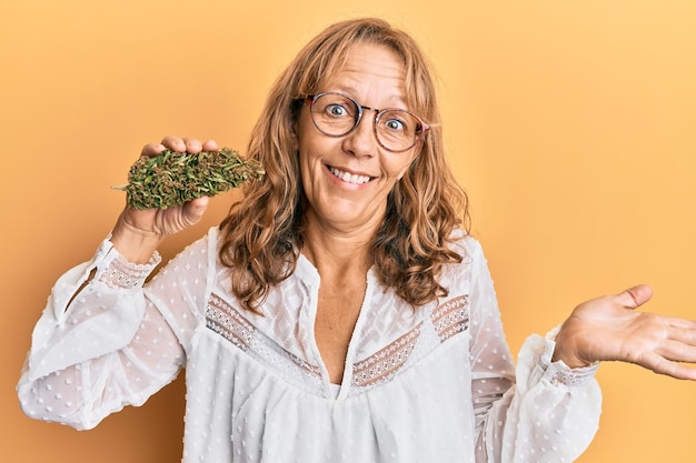 Free photo middle age blonde woman holding medical marijuana plant bud celebrating achievement with happy smile and winner expression with raised hand