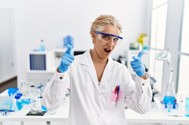 Бесплатное фото Блондинка средних лет, работающая в научной лаборатории, указывая пальцем на камеру со счастливым и забавным лицом, хорошей энергией и вибрациями
