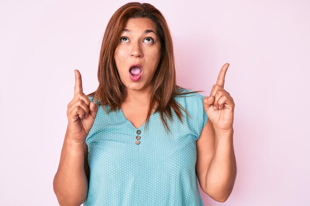 Middle age brunette hispanic woman wearing casual clothes amazed and surprised looking up and pointing with fingers and raised arms