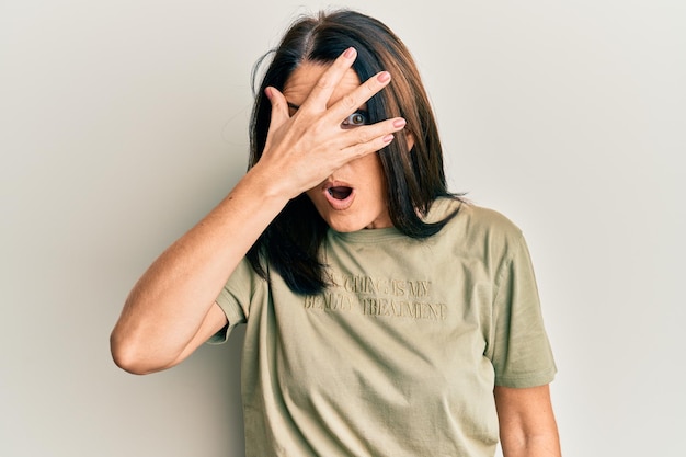 Middle age brunette woman wearing casual clothes peeking in shock covering face and eyes with hand, looking through fingers afraid