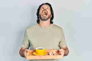 Free photo middle age caucasian man holding breakfast tray with cookies and coffee angry and mad screaming frustrated and furious shouting with anger looking up