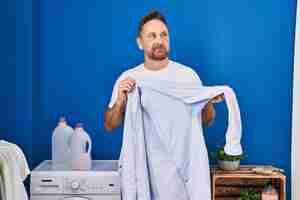 Free photo middle age caucasian man holding shirt with sweat smiling looking to the side and staring away thinking