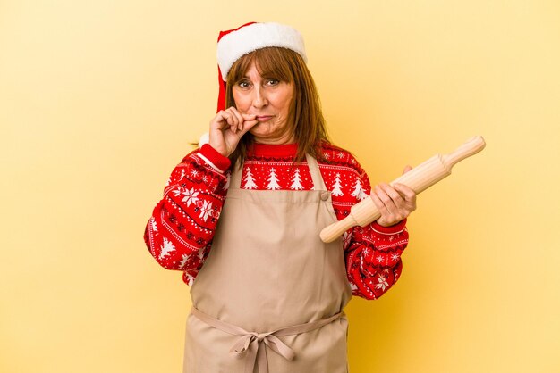 중년 백인 여성은 입술에 손가락을 대고 비밀을 유지하면서 노란색 배경에 격리된 크리스마스를 위해 쿠키를 요리합니다.