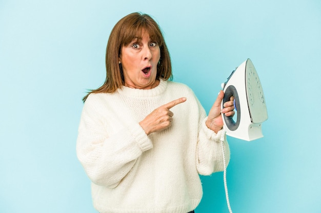 Middle age caucasian woman holding a iron isolated on blue background pointing to the side