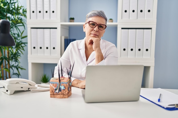 Middle age caucasian woman holding model of human anatomical skin and hair serious face thinking about question with hand on chin, thoughtful about confusing idea