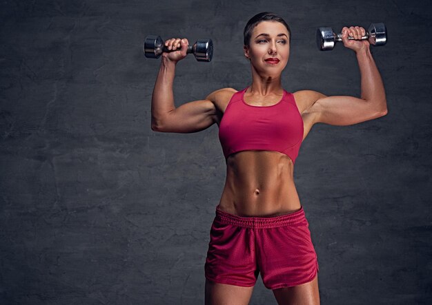 Middle age female with shirt hair holds dumbbells over grey background.