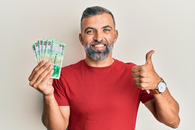 Middle age handsome man holding 50 hong kong dollars banknotes smiling happy and positive thumb up doing excellent and approval sign