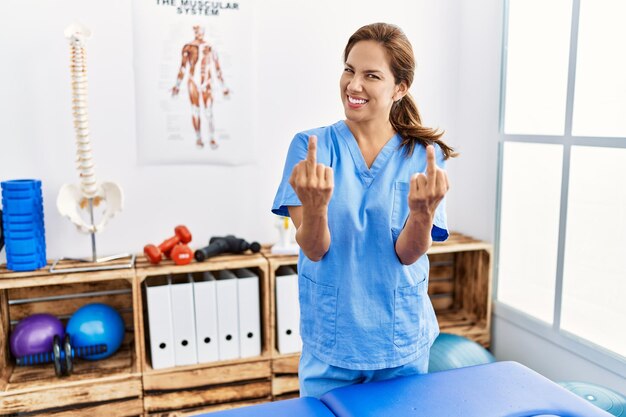 Middle age hispanic physiotherapist woman working at pain recovery clinic showing middle finger doing fuck you bad expression, provocation and rude attitude. screaming excited