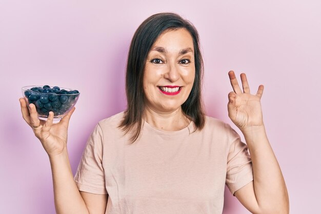 Middle age hispanic woman holding blueberries doing ok sign with fingers, smiling friendly gesturing excellent symbol