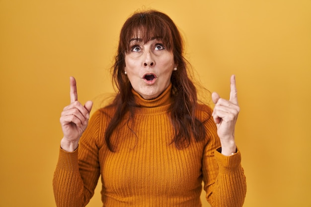 Middle age hispanic woman standing over yellow background amazed and surprised looking up and pointing with fingers and raised arms.