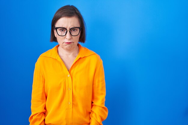 파란색 배경 위에 안경을 쓴 중년 히스패닉 여성은 회의적이고 긴장하며 문제 때문에 얼굴을 찌푸리고 있습니다. 부정적인 사람.