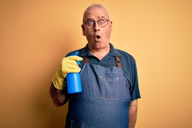 Пожилой уборщик средних лет чистит в фартуке и перчатках с помощью распылителя, испуганный в шоке с удивленным лицом, напуганный и взволнованный выражением страха