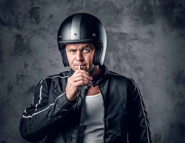 오토바이 헬멧과 가죽 재킷을 입은 중년 남성이 회색 배경에서 담배를 피우고 있습니다.