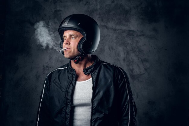 Мужчина средних лет в мотоциклетном шлеме и кожаной куртке курит сигарету на сером фоне.
