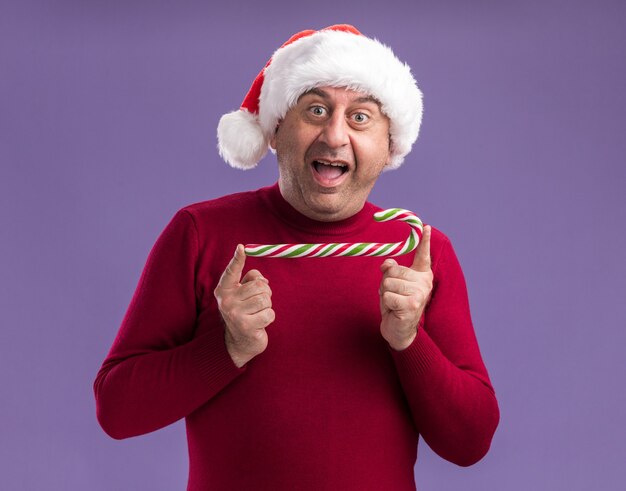 Middle age man wearing christmas santa hat holding candy cane looking at camera happy and excited standing over purple background
