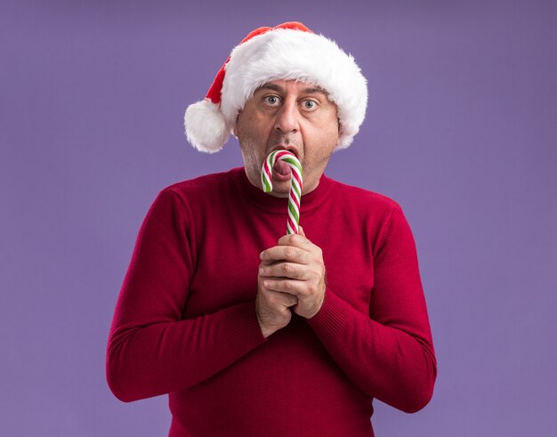 紫色の背景の上に立って驚いたカメラを見てキャンディケインを保持しているクリスマスサンタ帽子をかぶっている中年男性