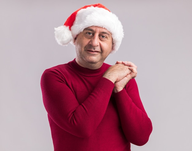 Middle age man wearing christmas santa hat  holding hands together  with smile on face  standing over white  wall