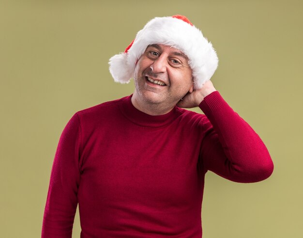 Foto gratuita uomo di mezza età che indossa il cappello di babbo natale che guarda l'obbiettivo sorridente felice ed emozionato allegramente in piedi su sfondo verde