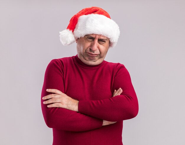 クリスマスのサンタの帽子をかぶった中年の男性は、白い壁の上に立って腕を組んで苦しそうな口を作って混乱して失望しているように見えます