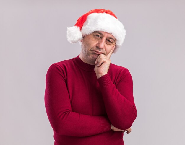 Middle age man wearing christmas santa hat  with skeptic expression  standing over white  wall