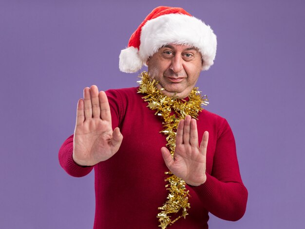 首に見掛け倒しのクリスマスサンタの帽子をかぶっている中年の男性は、紫色の壁の上に立って停止ジェスチャーを作る手を差し出すことに不満を持っています