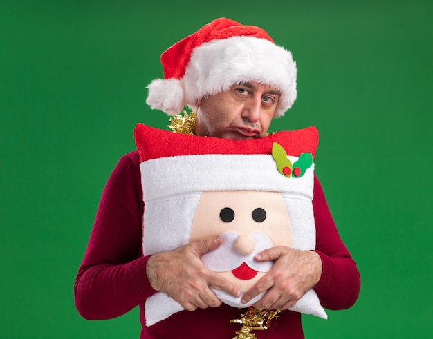 Middle age man wearing christmas santa hat with tinsel around neck holding christmas pillow looking at camera being displeased standing over green  background