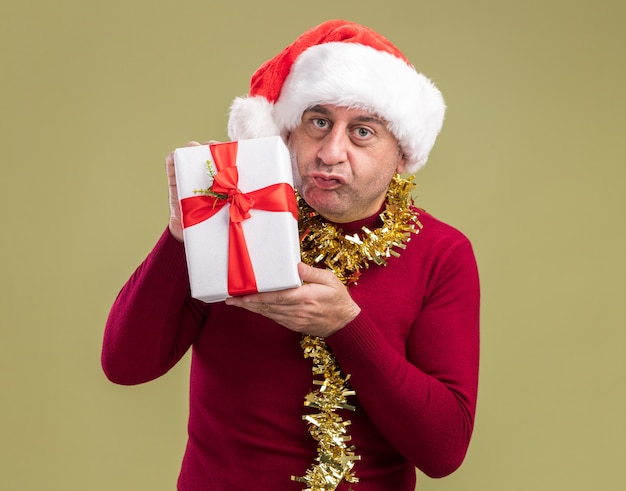 Uomo di mezza età che indossa il cappello di babbo natale con orpelli intorno al collo tenendo presente il regalo di natale guardando la fotocamera con espressione confusa in piedi su sfondo verde