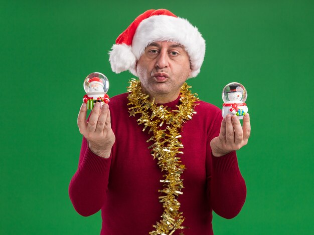 Middle age man wearing christmas santa hat with tinsel around neck holding christmas snow globes looking at camera with confident expression  standing over green  background