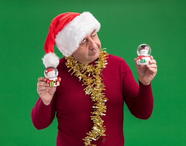 Middle age man wearing christmas santa hat with tinsel around neck holding christmas snow globes  looking confused having doubts  standing over green  background