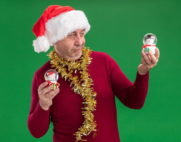 Middle age man wearing christmas santa hat with tinsel around neck holding christmas snow globes  looking confused trying to make choice 