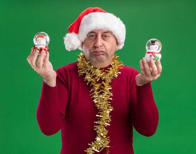 Free photo middle age man wearing christmas santa hat with tinsel around neck holding christmas snow globes   with skeptic expression