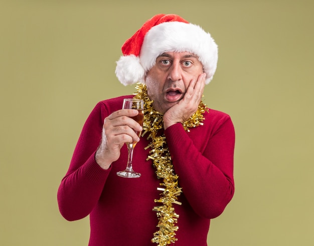 Middle age man wearing christmas santa hat with tinsel around neck holding glass of champagne  amazed and surprised standing over green  wall