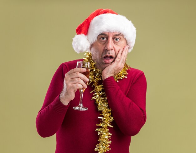 Мужчина средних лет в рождественской шапке санта-клауса с мишурой на шее с бокалом шампанского изумлен и удивлен, стоя у зеленой стены