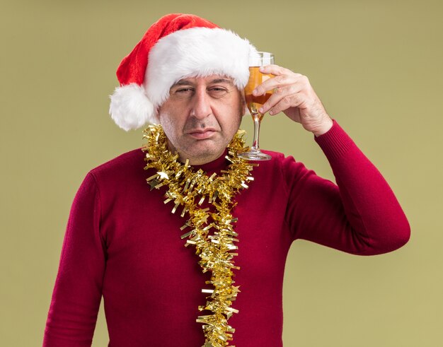 Middle age man wearing christmas santa hat with tinsel around neck holding glass of champagne  looking at camera with sad expression standing over green  background