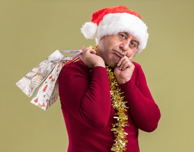 Мужчина средних лет в рождественской шляпе санта-клауса с мишурой на шее держит бумажные пакеты с рождественскими подарками, уставший и скучающий, глядя в камеру, стоя на зеленом фоне