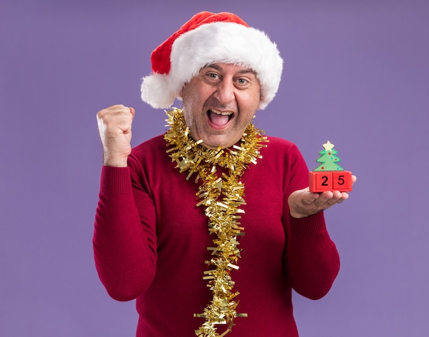 Middle age man wearing christmas santa hat  with tinsel around neck holding toy cubes with date twenty five happy and excited clenching fist standing over purple background