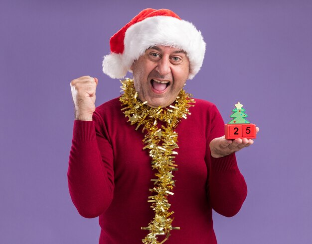 보라색 배경 위에 서있는 날짜 25 행복하고 흥분 떨림 주먹으로 장난감 큐브를 들고 목 주위에 반짝이와 크리스마스 산타 모자를 쓰고 중년 남자