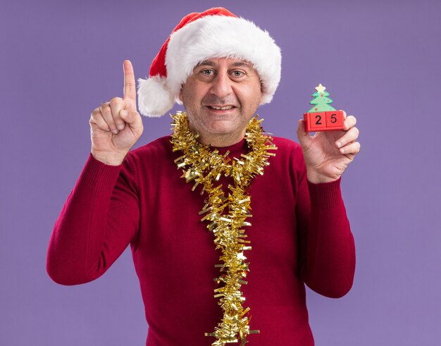 Middle age man wearing christmas santa hat  with tinsel around neck holding toy cubes with date twenty five  looking at camera smiling showing index finger standing over purple background