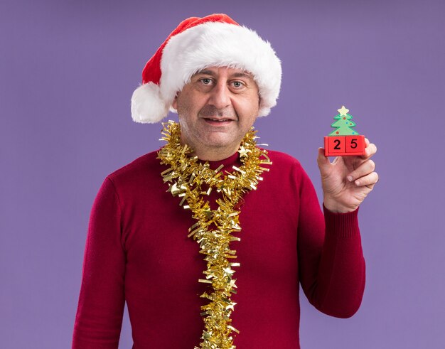 мужчина среднего возраста в рождественской шляпе санта-клауса с мишурой на шее держит игрушечные кубики с датой двадцать пять, глядя в камеру, улыбаясь, стоя на фиолетовом фоне