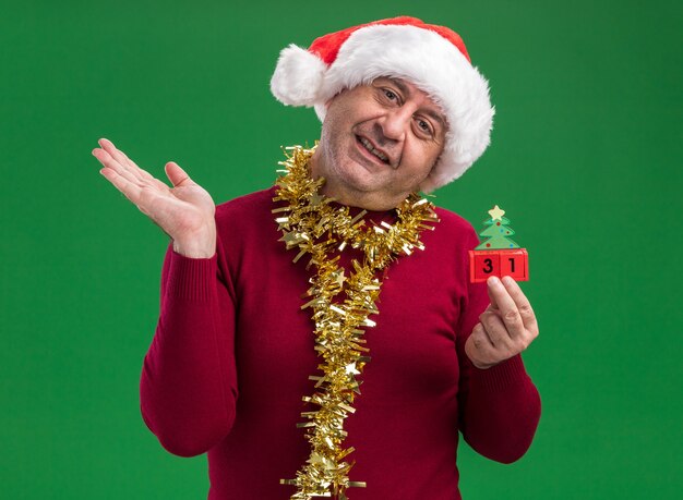 Мужчина средних лет в рождественской шапке санта-клауса с мишурой на шее держит игрушечные кубики с новогодней датой, глядя в камеру, улыбаясь со счастливым лицом, поднимая руку, стоя на зеленом фоне