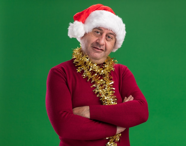 Middle age man wearing christmas santa hat with tinsel around neck  looking at camera  smiling confident with arms crossed standing over green  background