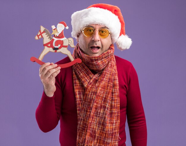 紫色の背景の上に立って驚いたカメラを見てクリスマスのおもちゃを保持している黄色いメガネで首の周りに暖かいスカーフとクリスマスサンタ帽子をかぶっている中年男性