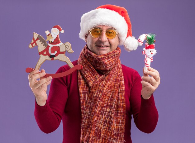 Middle age man wearing christmas santa hat with warm scarf around neck  in yellow glasses showing christmas toys happy and cheerful smiling  standing over purple background