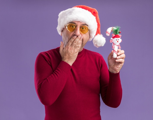 Foto gratuita uomo di mezza età che indossa il cappello della santa di natale in vetri gialli che tengono il bastoncino di zucchero di natale che guarda l'obbiettivo stupito che copre la bocca con la mano che sta sopra il fondo viola