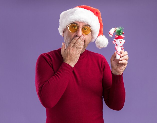 카메라를보고 크리스마스 사탕 지팡이를 들고 노란색 안경에 크리스마스 산타 모자를 쓰고 중년 남자는 보라색 배경 위에 서있는 손으로 입을 덮고 놀랐습니다.