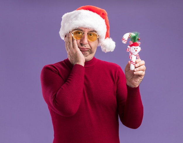 Middle age man wearing christmas santa hat in yellow glasses holding christmas candy cane looking at camera confused standing over purple background