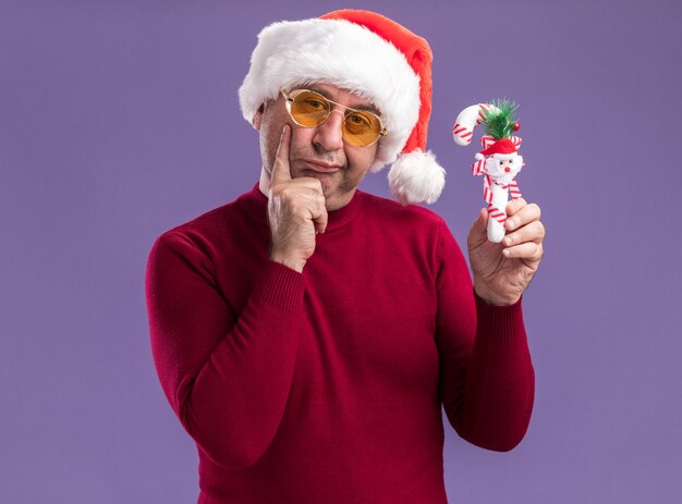 Мужчина средних лет в рождественской шляпе санта-клауса в желтых очках держит рождественскую конфету, глядя в камеру со скептическим выражением лица, стоя на фиолетовом фоне