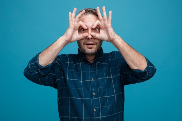 Uomo di mezza età con capelli grigi in camicia blu scuro che guarda la fotocamera attraverso le dita che fanno un gesto binoculare in piedi su sfondo blu