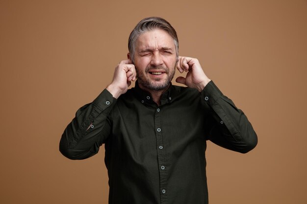 Middle age man with grey hair in dark color shirt closing ears with fingers with annoyed expression standing over brown background